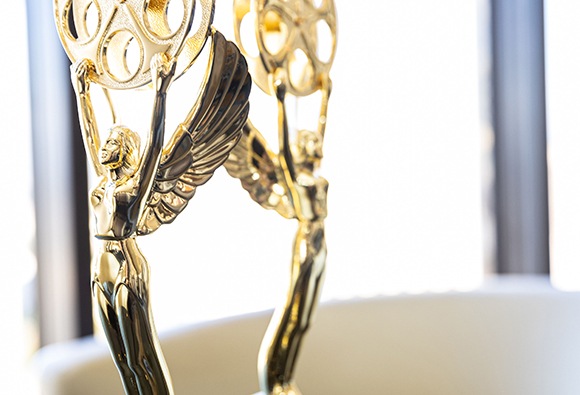 Two golden winged statues holding film reels against a bright, blurred background.