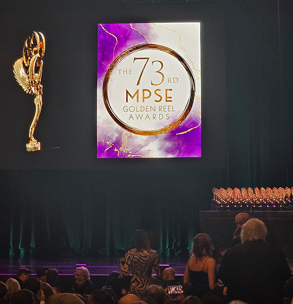A golden trophy stands to the left of a screen displaying the 73rd MPSE Golden Reel Awards logo. An audience is seated in the