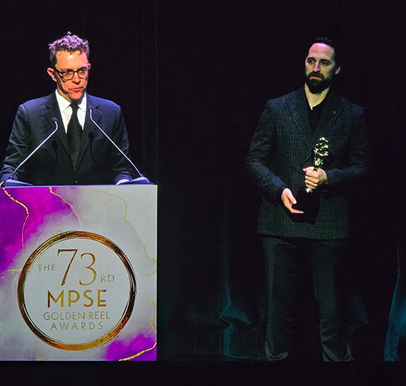 A man in a suit stands at a podium with the 73rd MPSE Golden Reel Awards logo. Another man holding an award stands to his rig