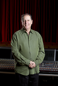 A smiling man wearing a green button-down shirt stands in front of a red curtain and a mixing console.