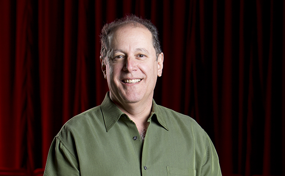 A smiling man in a green collared shirt posing in front of a dark red curtain.