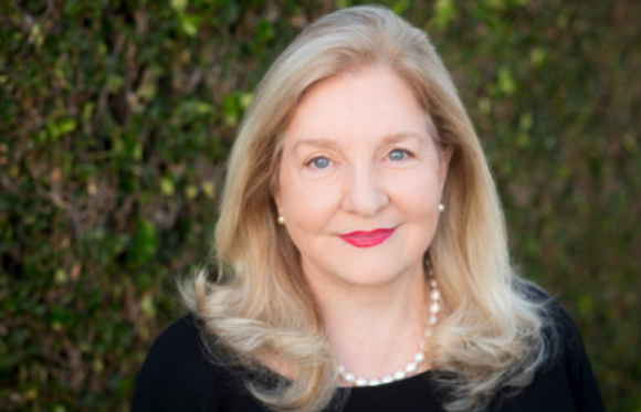 A blonde woman with pearl earrings and a pearl necklace smiles in front of a green hedge.