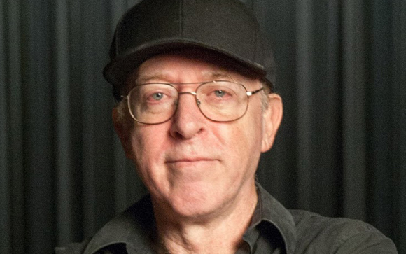 A man with glasses and a black baseball cap looking directly at the camera against a dark curtain.