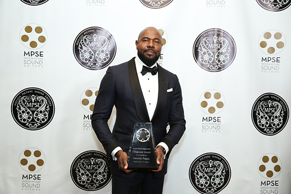 A man in a black tuxedo holding a dark award in front of a white backdrop with MPSE logos.
