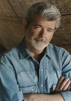 A man with wavy gray hair and a gray beard wearing a blue denim shirt, looking at the camera with arms crossed.