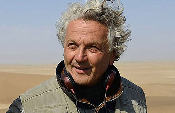 A smiling man with curly grey hair wearing a vest and headphones in a desert landscape.