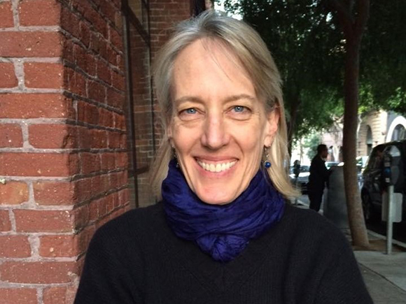 A smiling woman with blonde hair and a blue scarf standing outdoors near a brick wall.