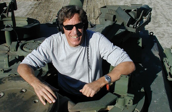 Smiling man wearing sunglasses and headphones sitting inside an open hatch of a green military vehicle.