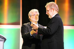 Two men in formal suits stand on a stage with a colorful background, one holding a small object.