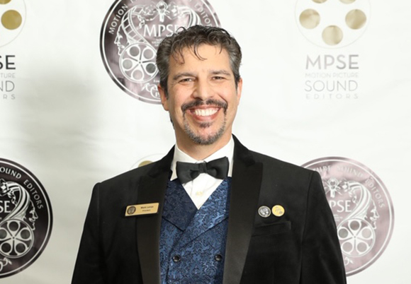 A man with dark hair and a mustache wearing a black tuxedo and blue patterned vest, smiling at the camera.