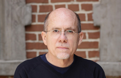 A man with glasses and a goatee looking directly at the camera against a brick background.