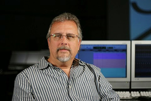 A man with graying hair and glasses wearing a striped shirt, sitting in a studio with computer monitors.
