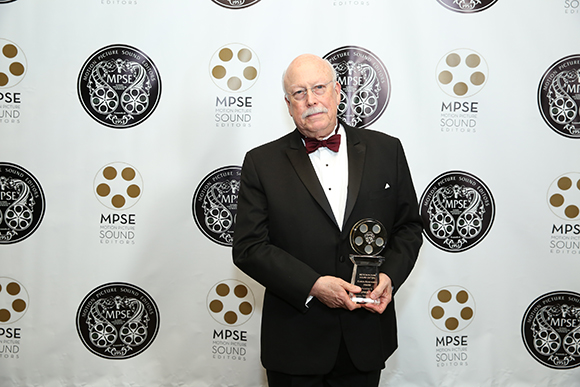 A man with white hair and a mustache wearing a tuxedo and bowtie, holding a glass award.