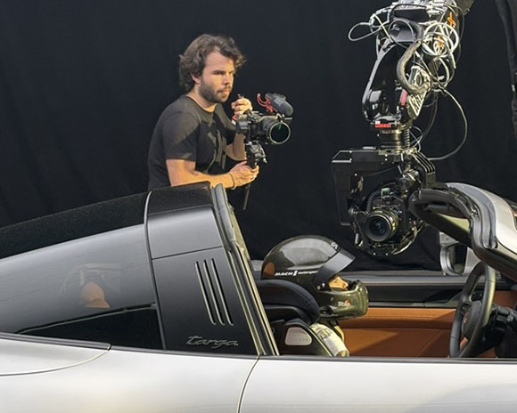A man holding a professional camera films a person wearing a helmet inside a silver convertible car.