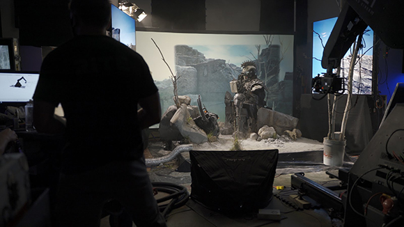 A person in silhouette filming a studio set featuring a large screen with a landscape and rocky terrain.