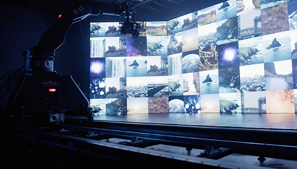 A robotic camera arm in a dark studio facing a massive wall of screens showing diverse landscape imagery.