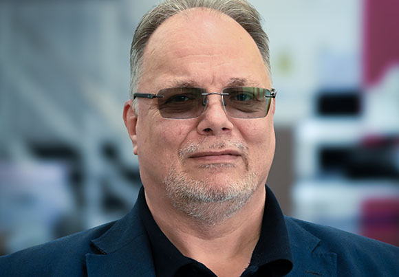 A man with light skin, short hair, and glasses wearing a dark blue blazer over a black shirt.