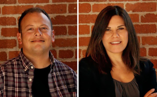 A man in a plaid shirt and a woman in a black blazer pose against a red brick background.