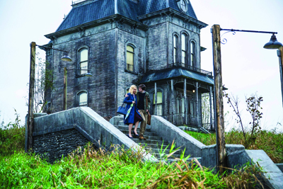 A woman in a blue dress stands on stone steps leading up to a large, weathered wooden house.