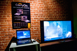 A poster for CarryOn hangs above a laptop and a large monitor in a dark room with a brick wall.