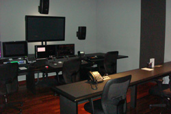A dark editing studio with multiple monitors, speakers, and black desks with office chairs.