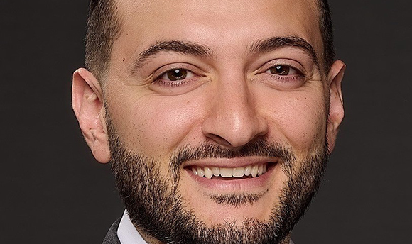 A smiling man with dark hair, brown eyes, and a groomed beard against a dark background.