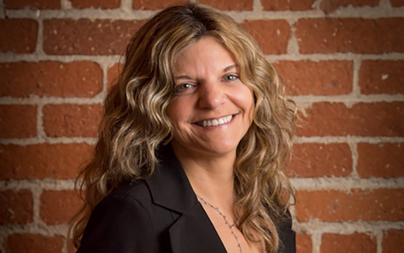 A smiling woman with long, wavy blonde hair wearing a black blazer against a red brick wall.
