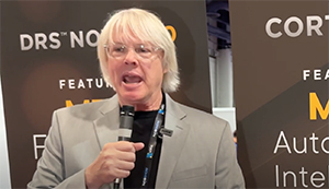 A man with white hair and glasses speaks into a handheld microphone in front of a branded backdrop.