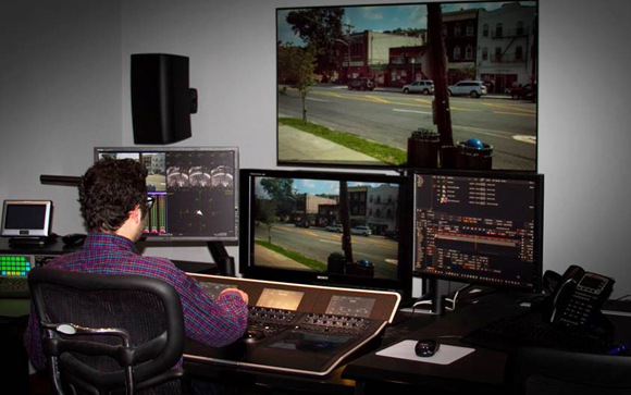 A person in a plaid shirt sits at a desk with four monitors displaying video editing software and footage.