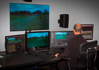 A person sits at a desk with four computer monitors displaying video editing software and footage.