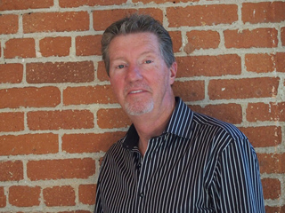 A man with short brown hair wearing a black and white striped shirt against a red brick wall.