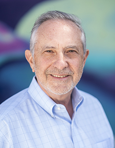 A smiling man with graying hair and a short beard wearing a light blue checkered button-down shirt.