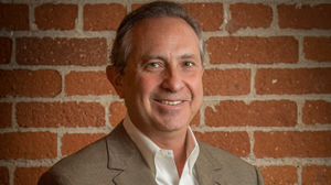 A smiling man in a tan blazer and white shirt posing in front of a red brick wall.