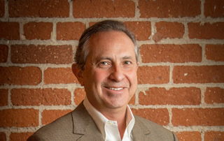 A smiling man wearing a tan blazer and white collared shirt in front of a red brick wall.