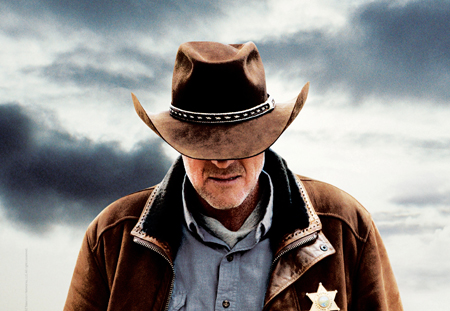 A man wearing a brown cowboy hat and a brown jacket with a sheriff's star badge looking downward.