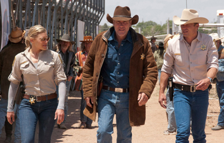 Several people wearing cowboy hats and sheriff badges walk along a dirt path in a sunny outdoor setting.