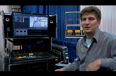 A man in a grey shirt sits beside a large media server rack with a monitor and control panel.