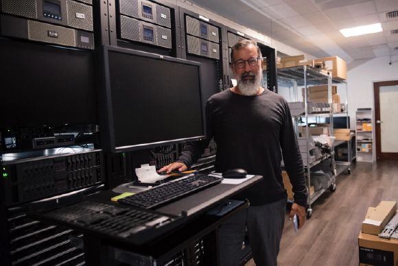 A man with a grey beard stands by a computer monitor and keyboard in a room filled with server racks.