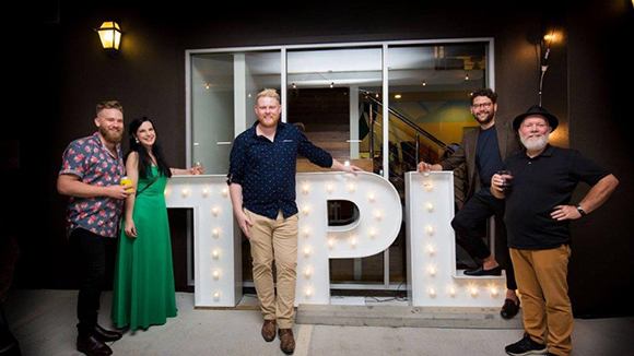 Five people standing behind large, lit-up letters spelling TPL at night.