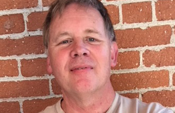 A man with short brown hair smiling in front of a red brick wall.