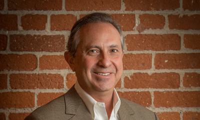 A smiling man in a tan blazer and white shirt poses in front of a red brick wall.
