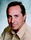 A man with dark hair and a tan collared shirt looking toward the camera against a light blue background.