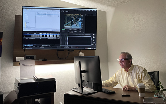 A man in a yellow shirt sits at a desk looking at a computer monitor in an office setting.
