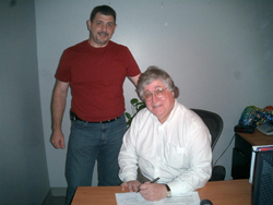 A man in a red t-shirt stands next to a man in a white shirt sitting at a desk.