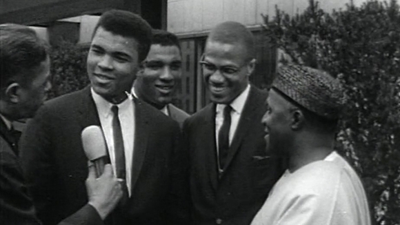 A young Muhammad Ali in a suit is interviewed by men outdoors near some bushes.