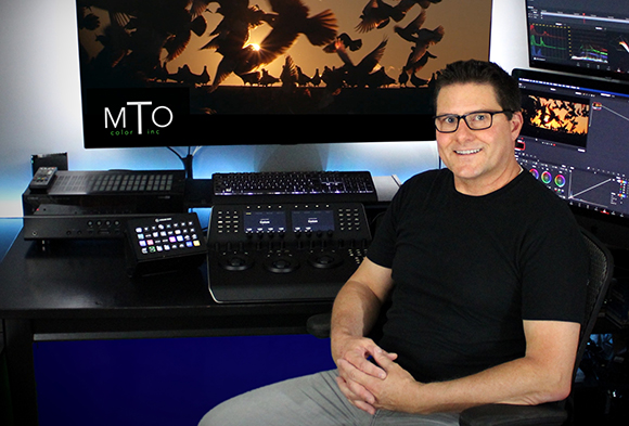 A man in a black t-shirt sits at a desk with multiple monitors and color grading control panels.
