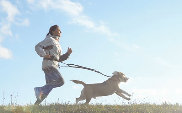 A woman runs with her dog on a leash across a grassy field under a bright blue sky with scattered clouds.