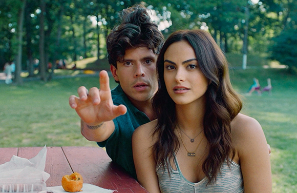 A young man reaches toward the camera while a young woman looks forward at an outdoor table.