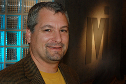 A smiling man with short graying hair wearing a brown jacket and yellow shirt.