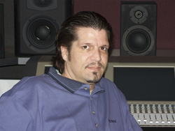 A man with dark hair and a goatee sitting in front of studio monitors and a mixing console.